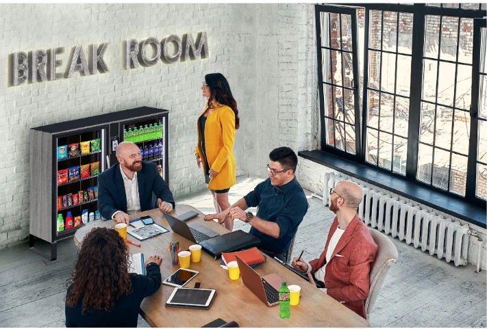 A modern break room with a snack-filled cabinet and a diverse group of professionals engaged in discussion around a table.